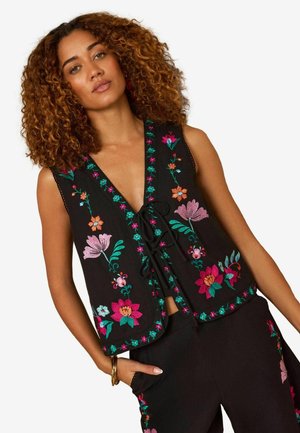 Frau mit lockigem Haar trägt eine schwarze ärmellose Weste und Hose, beide mit bunter Blumenstickerei verziert.