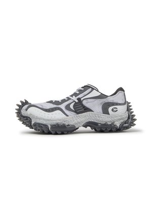 Chaussure de sport blanche et grise avec empeigne en mesh texturé et crampons en caoutchouc noir dépassant de la semelle et du talon.