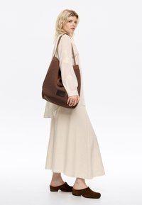 Borsa tote in tonalità marrone con manico a taglio circolare, indossata su una camicia lunga a maniche fluente di colore crema e un completo gonna.