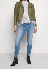 Zielona oliwkowa kurtka motocyklowa z zamszu z srebrnymi elementami, zestawiona z białym i granatowym paskowanym T-shirtem oraz jasnoniebieskimi obcisłymi dżinsami. Czarne botki za kostkę.