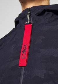 Mørkeblå jakke med camouflage mønster, udstyret med lynlås lukning, en rød stof dragetab og et logo på tabben. Synligt tekstureret stof detaljer.