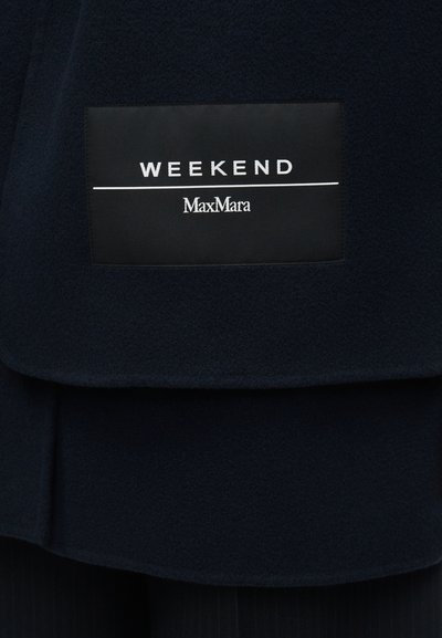 Μαύρη ορθογώνια ετικέτα ρούχου με λευκό κείμενο "WEEKEND" πάνω από μια γραμμή και "MaxMara" κάτω σε σκούρο ύφασμα.