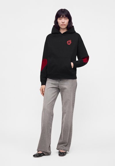 Femme portant un sweat à capuche noir avec des patches rouges aux coudes et le logo Apple, un jean gris clair et des ballerines noires, debout devant un fond uni.