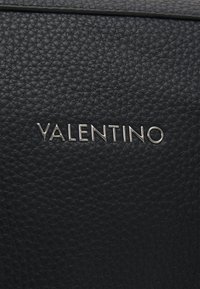 Svart lädermaterial med en texturerad yta. Silverfärgad "VALENTINO"-logotyp som är tydligt placerad i mitten.
