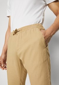 Beige elastic waist pants with an adjustable drawstring, featuring side pockets and a smooth, lightweight fabric.