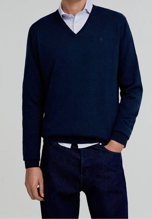 Homme portant un pull bleu marine à col en V par-dessus une chemise bleu clair boutonnée et un jean foncé, une main dans la poche sur fond uni.
