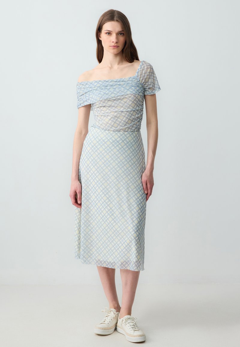 Off-shoulder jurk met een blauw en crèmekleurig geruit patroon, doorschijnende textuur en een aansluitend lijfje dat overgaat in een vloeiende rok.