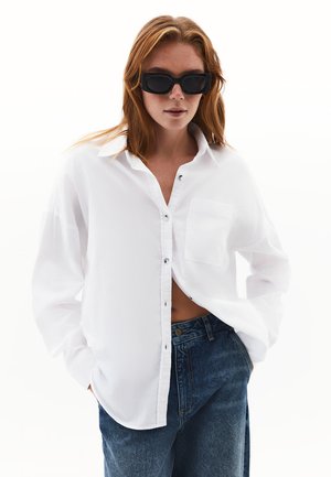 Camicia bianca con bottoni, dotata di taschino, vestibilità rilassata e maniche lunghe, abbinata a jeans in denim blu a vita alta. Occhiali da sole neri completano il look.
