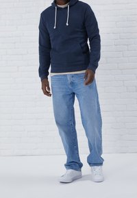 Marinblå hoodie med justerbara dragkedjor, framficka; ljusblåjeans; vita sneakers. Avslappnad outfit mot en vit tegelvägg.