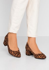 Leopard print high-heeled shoes with a textured suede finish, a square toe design, and a chunky heel, paired with light-colored trousers.