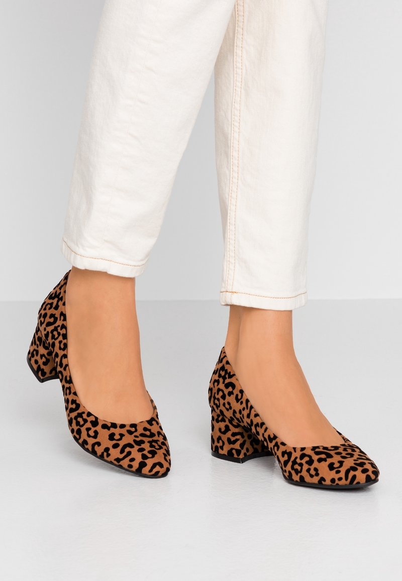 Leopard print high-heeled shoes with a textured suede finish, a square toe design, and a chunky heel, paired with light-colored trousers.