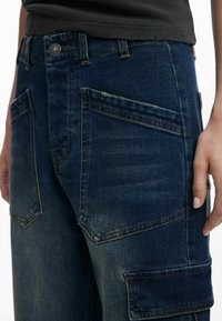 Pantalones cargo de mezclilla en azul oscuro, con una cintura alta, múltiples bolsillos, costuras visibles y una textura desgastada en la tela.