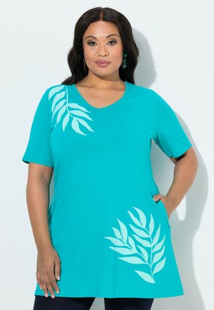 Mujer de cabello largo y oscuro, vestida con una túnica verde azulado con patrones de hojas en azul claro, posando con una mano en la cadera contra un fondo blanco.