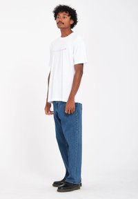 White T-shirt with pink graphic on the front, paired with loose blue jeans and black shoes. Plain background with natural lighting.