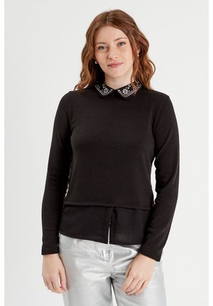Haut à manches longues noir avec un col orné contrasté. Présente une coupe ample et un détail fendu à l'ourlet. Associé à un pantalon argenté brillant.