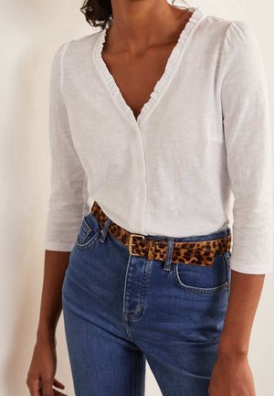 Woman wearing white ruffle-edge buttoned top with three-quarter sleeves and blue jeans with leopard print belt and gold buckle.