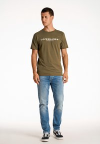 Camiseta verde oliva con el texto "COPENHAGUE LINDBERGH", combinada con jeans de mezclilla azul claro y zapatillas en blanco y negro. Ajuste casual, mangas cortas.
