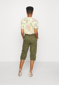 Květinová košile s krátkým rukávem v pastelově žluté barvě s zelenými a růžovými květinovými vzory, doplněná olivově zelenými zkrácenými kalhotami a bílými sandály na podpatku.