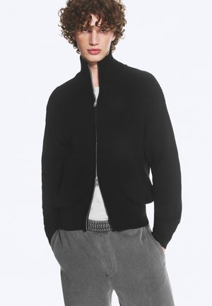 Jeune personne aux cheveux bouclés portant un pull noir zippé sur une chemise blanche et un pantalon gris décontracté, les mains dans les poches, debout sur fond blanc.