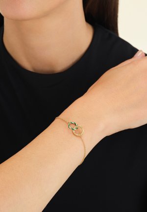 Pulsera de cadena de oro con anillos entrelazados, uno de diseño dorado y otro de diseño verde retorcido, llevada en una muñeca sobre un fondo negro.
