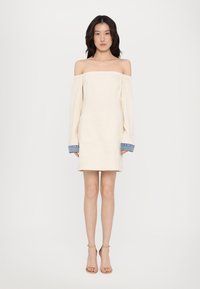 Rochie bej off-the-shoulder, cu mâneci lungi, manșete din denim și o siluetă dreaptă. Textură netedă și un design casual.