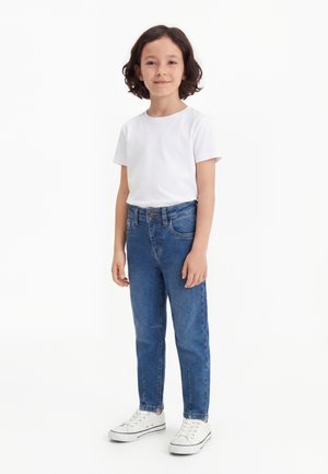 Enfant aux cheveux foncés mi-longs portant un t-shirt blanc, un jean bleu et des baskets blanches, debout devant un fond blanc uni.