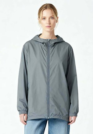 Vrouw draagt een grijze capuchonvest met rits en elastische manchetten, gecombineerd met lichtblauwe spijkerbroek, tegen een effen lichte achtergrond.