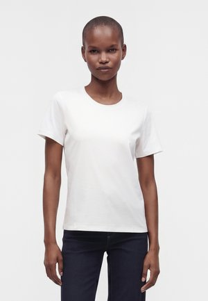 Vrouw met kort haar die een eenvoudig wit T-shirt met korte mouwen en donkere spijkerbroek draagt, staand voor een effen lichte achtergrond.