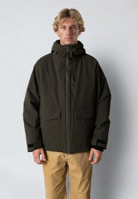 Tumma vihreä vedenpitävä takki, jossa on korkea kaulus, etuvetoketju, kaksi suurta taskua ja säädettävät hihansuut. Rakennekangas ja huppu.