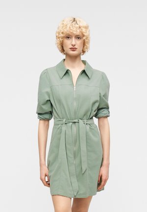 Kvinde med krøllet blondt hår iført en lysegrøn kjole med bælte, opkrogede ærmer og lynlås foran, stående mod en ensfarvet baggrund.