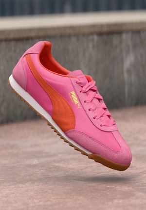 Różowo-pomarańczowy sneaker Puma z sznurowadłami, białą podeszwą i teksturowanym brązowym bieżnikiem na rozmytej, zewnętrznej powierzchni.