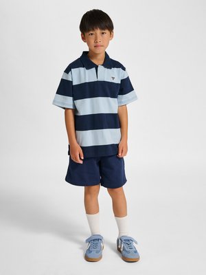 Dreng stående iført navyblå og lyseblå stribet polo, navyblå shorts, hvide sokker og lyseblå sneakers mod ensfarvet baggrund.
