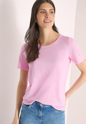 Kvinde med langt mørkt hår iført en lyserød kortærmet skjorte og blå jeans, stående med den ene hånd på hoften.