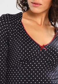 Haut marine avec des pois blancs, présentant un décolleté en V bordé de rouge et une petite accent en forme de nœud. Le tissu semble doux avec une texture lisse.