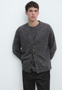 Mörkgrå ullkofta med V-ringning, långa ärmar och ett strukturerat stickmönster, med fyra knappar framtill. Bärs över en mörk skjorta.