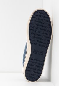 Modré plátěné tenisky s texturovanou podrážkou, mají světle béžový lem a reliéfní branding "TOMMY JEANS" na spodní straně.