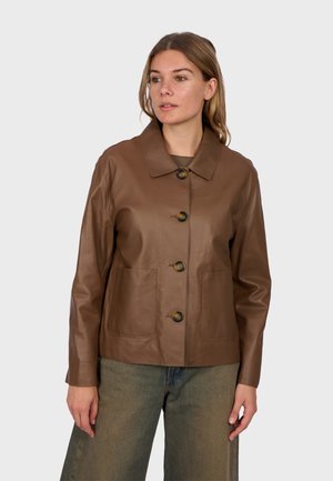 Frau trägt eine braune lederne Jacke mit Knöpfen und zwei vorderen Taschen sowie grünlich-graue weite Hosen und schaut leicht nach links.