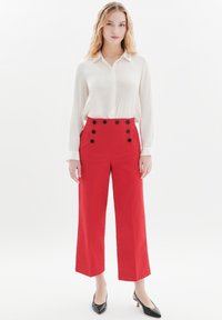 Röda vida byxor med hög midja, med en dubbelknäppt design och sex svarta knappar, parat med en vit långärmad blus.