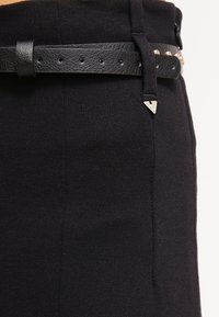 Zwarte high-waisted broek van een gestructureerde stof. Voorzien van een zwarte leren riem met gouden studs en een metalen driehoekaccent.