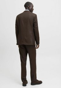 Braun maßgeschneiderter Anzug mit spitzem Revers, einem hinteren Schlitz und passender Hose. Weiche Stoffstruktur mit sichtbaren Nähdetails.