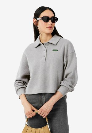 Femme aux longs cheveux foncés porte des lunettes de soleil noires, un polo gris à manches longues Lacoste, un jean sombre, et tient un sac à main beige devant elle.