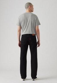 Muškarac stoji okrenut leđima, odjeven u sivi majicu, crne traperice ravnog kroja i crno-bijele tenisice, protiv jednostavne pozadine.