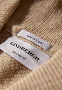Beżowa tkanina z lambswool z teksturowanym splotem. Metki z napisem "LINDNERGH" i "RELAXED FIT" w czarnym kolorze, oraz "LAMBS WOOL" w białym.