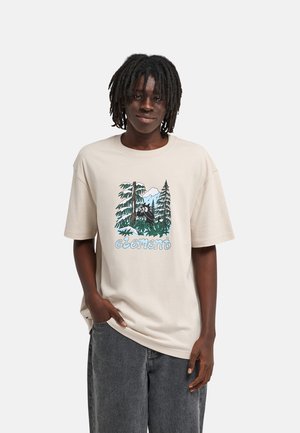 Personne avec des dreadlocks portant un t-shirt beige avec un motif forêt et le mot "element,", associé à un pantalon ample gris foncé.