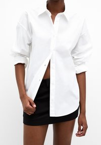 Chemise blanche à manches longues avec texture de tissu visible, dotée d'un col et d'un ourlet arrondi, associée à une mini-jupe noire.
