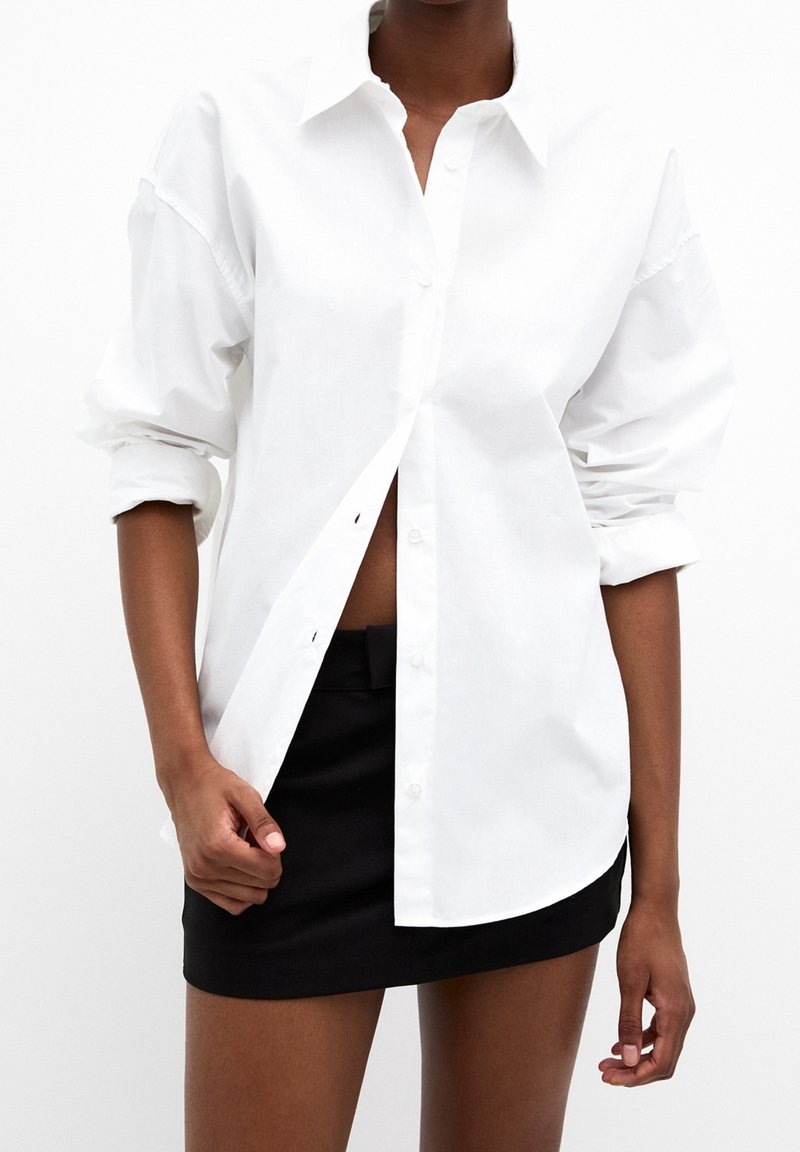 Chemise blanche à manches longues avec texture de tissu visible, dotée d'un col et d'un ourlet arrondi, associée à une mini-jupe noire.