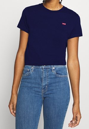 Kvinde iført en mørkeblå Levi's t-shirt og højtaljede lyseblå jeans står mod en ensfarvet baggrund.