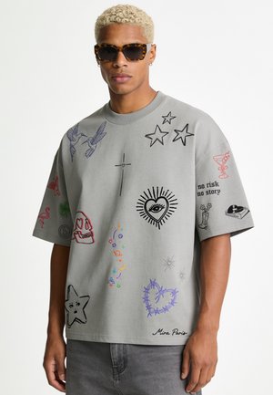 Homme aux cheveux blonds bouclés portant des lunettes de soleil écaille et un t-shirt gris oversize avec des gribouillis graphiques colorés et diverses petites illustrations.