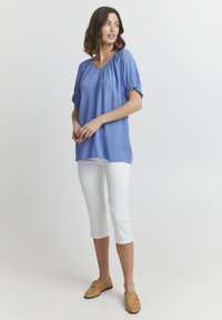 Blusa azzurra chiara con maniche a palloncino corte, scollatura a V e texture morbida, abbinata a pantaloni capri bianchi e mocassini beige.