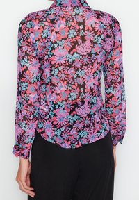 Blusa floral de chifón transparente, con flores rosas y azules sobre un fondo negro, mangas largas y dobladillo curvado.